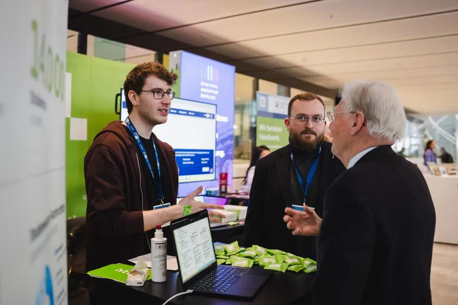 Mitarbeiter erklärt zwei Gästen am Mini-Expo-Stand ein digitales Werkzeug.