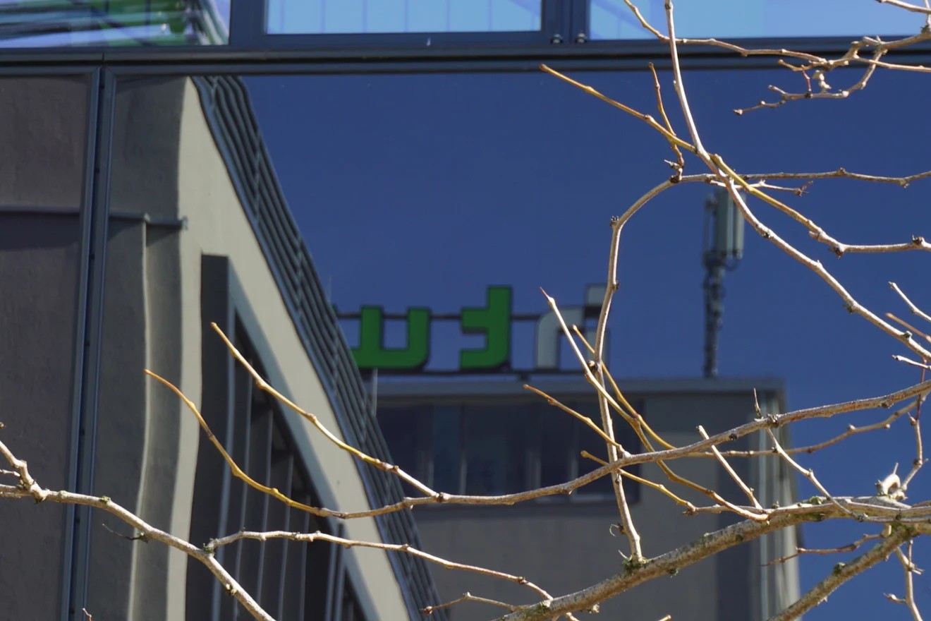 HTW-Logo an der Glasfassade am Campus Wilhelminenhof, hinter kahlen Zweigen.